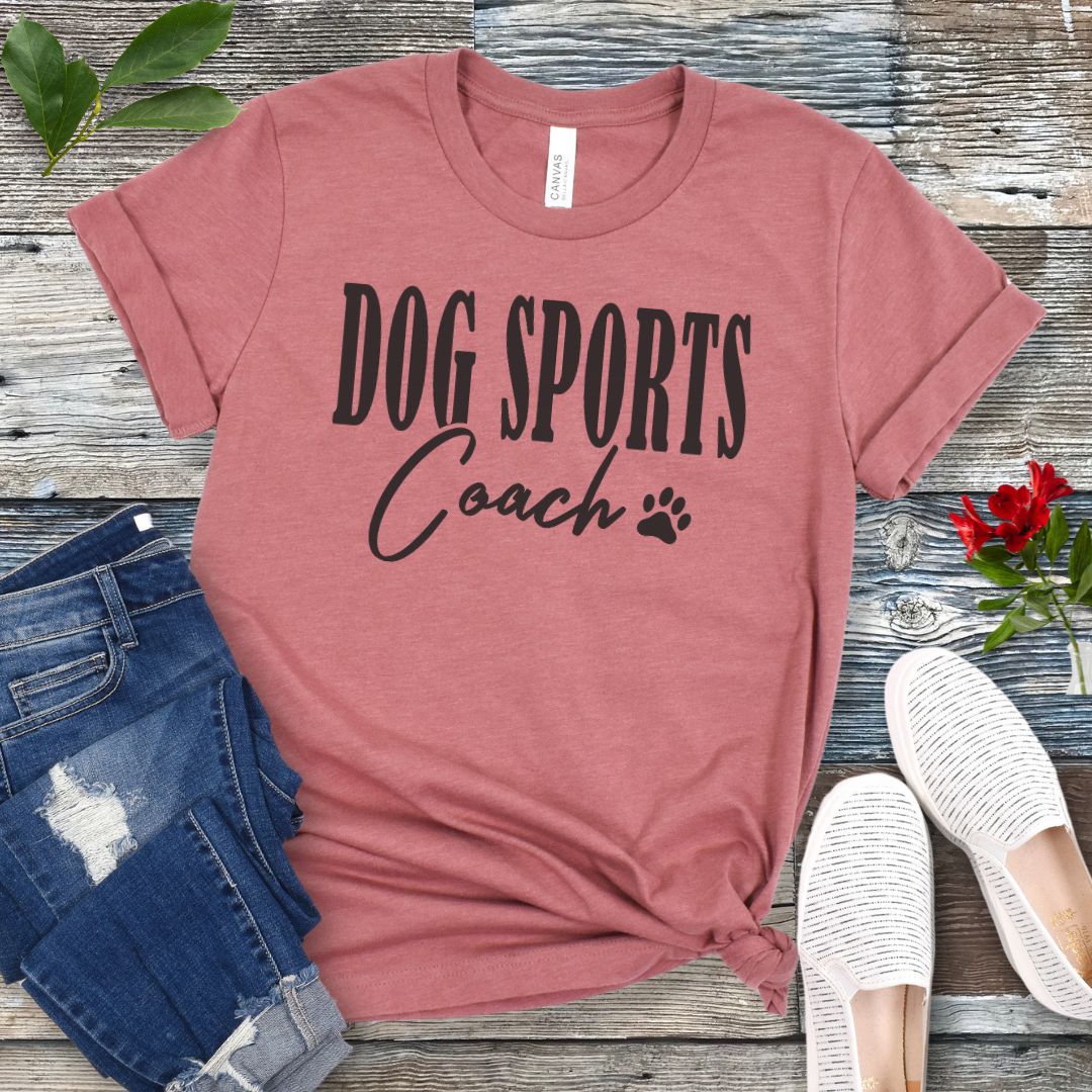 Mauve t-shirt with 'DOG SPORTS Coach' text and a paw print on a wooden surface with jeans and shoes.