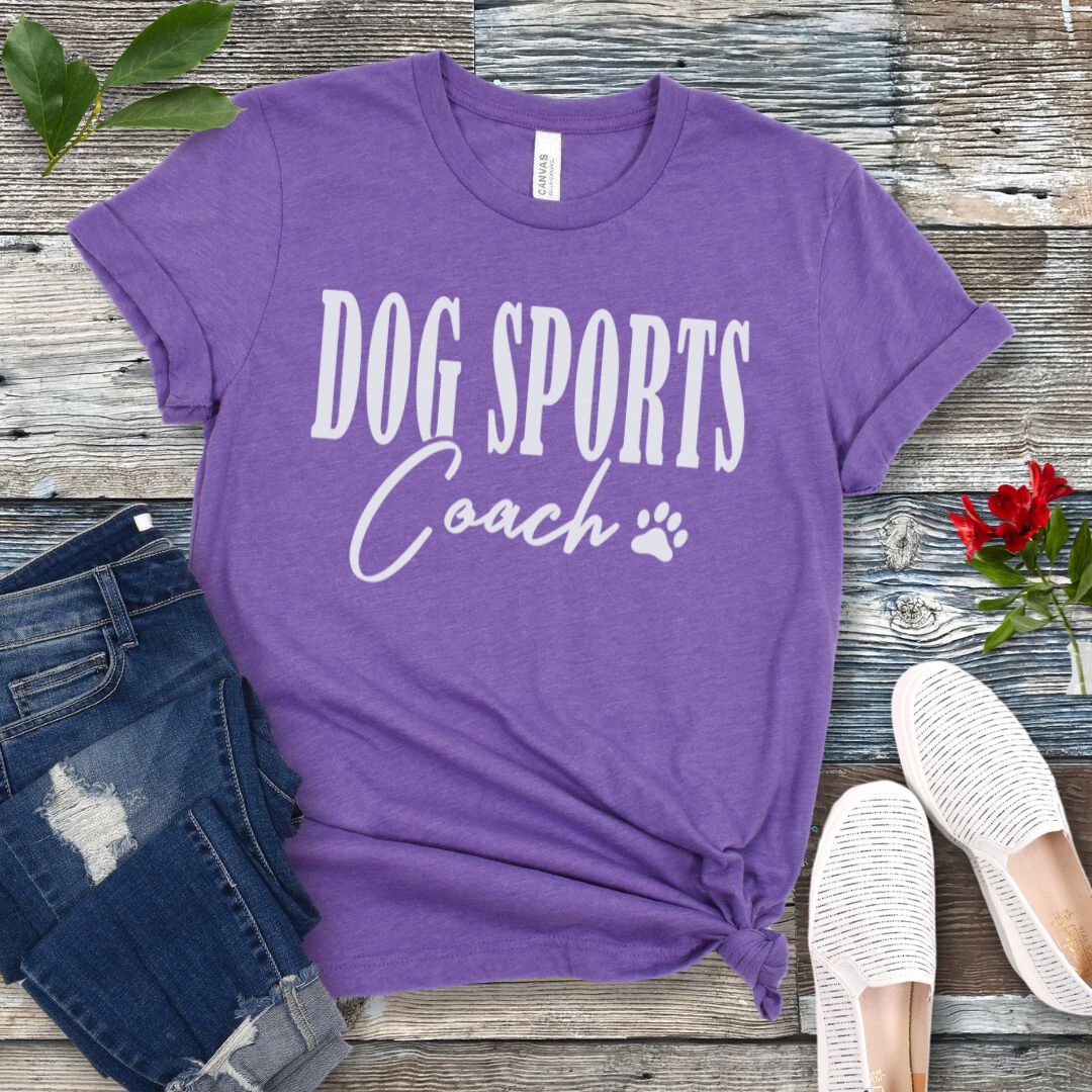 Purple t-shirt with 'DOG SPORTS Coach' text and a paw print on a wooden surface with jeans and shoes.
