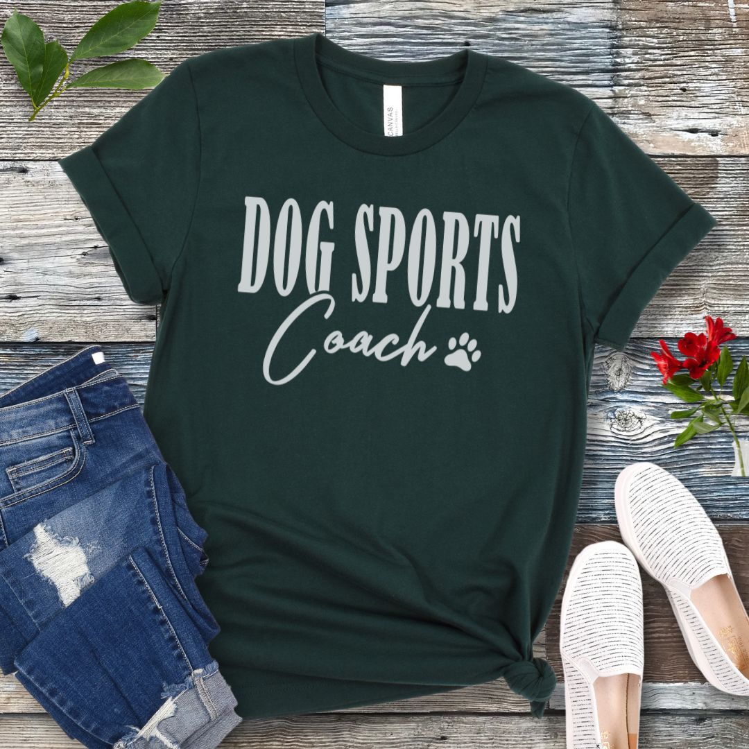 Dark green t-shirt with 'DOG SPORTS Coach' text and a paw print on a wooden surface with jeans and shoes.