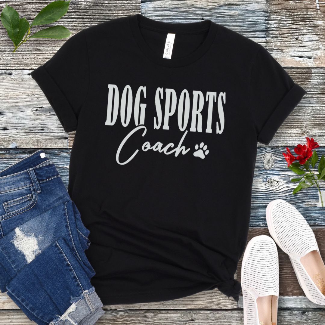 Black t-shirt with 'DOG SPORTS Coach' text and a paw print on a wooden surface with jeans and shoes.