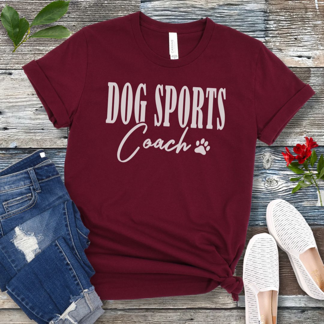 Maroon t-shirt with 'DOG SPORTS Coach' text and a paw print on a wooden surface with jeans and shoes.