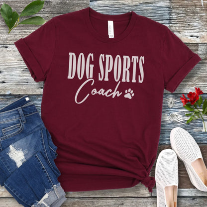 Maroon t-shirt with 'DOG SPORTS Coach' text and a paw print on a wooden surface with jeans and shoes.