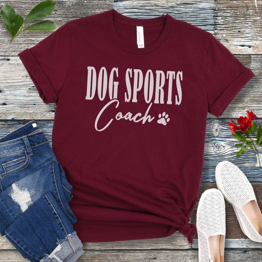 Maroon t-shirt with 'DOG SPORTS Coach' text and a paw print on a wooden surface with jeans and shoes.