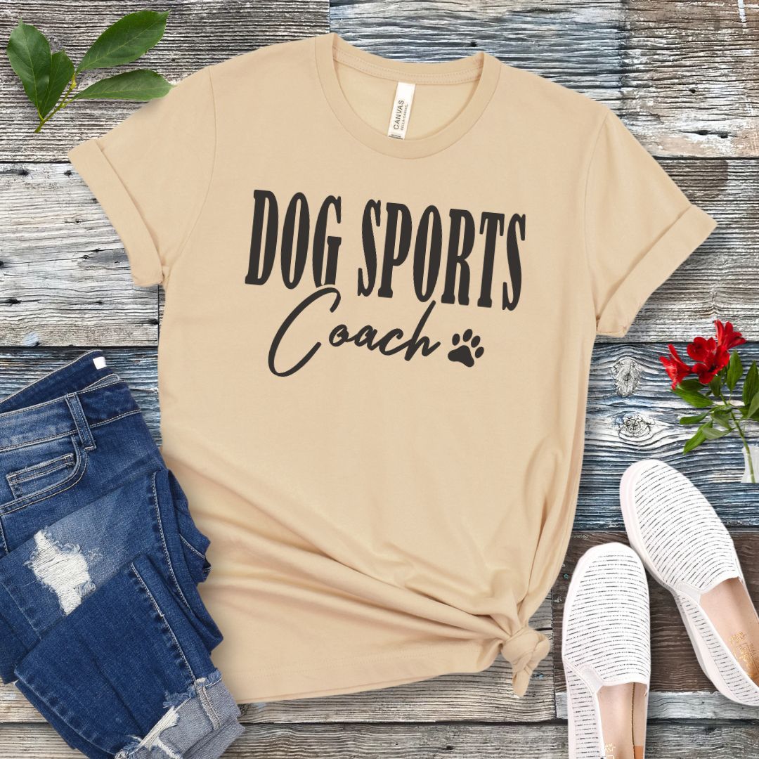 Cream colored t-shirt with 'DOG SPORTS Coach' text and a paw print on a wooden surface with jeans and shoes.