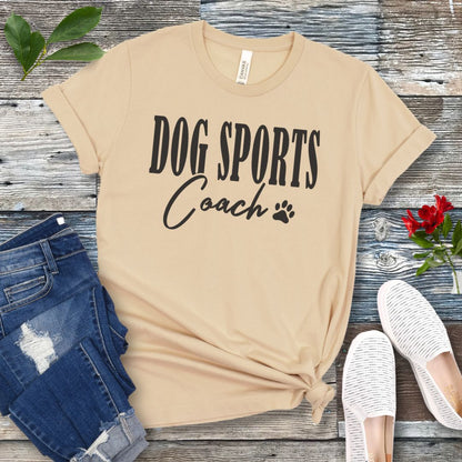 Cream colored t-shirt with 'DOG SPORTS Coach' text and a paw print on a wooden surface with jeans and shoes.