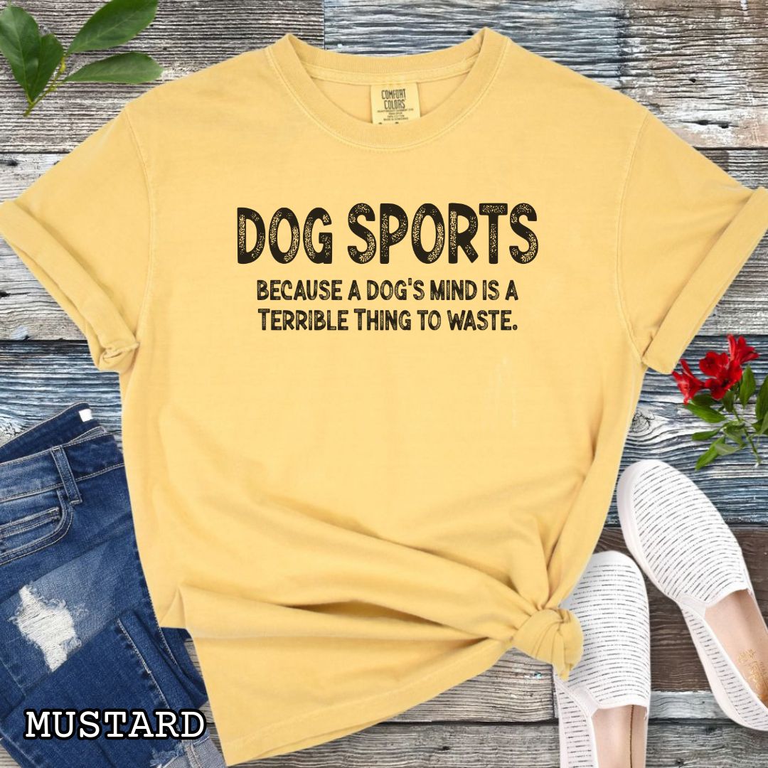 Mustard yellow Comfort Colors t-shirt for agility, nosework, obedience, flyball and more, with 'Dog Sports, because a dog's mind is a terrible thing to waste' text, shown on a wooden surface with shoes and jeans.