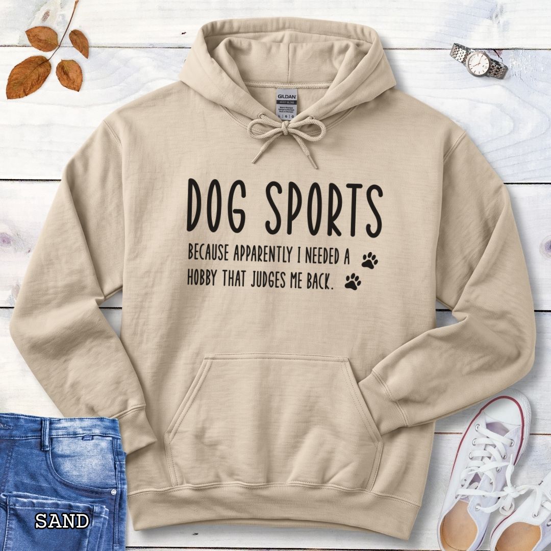 Beige unisex hoodie for dog training enthusiasts with humorous text 'Dog Sports, Because Apparently I Needed a Hobby That Judges Me Back' accompanied by paw prints, shown on a wooden surface with jeans and flowers.