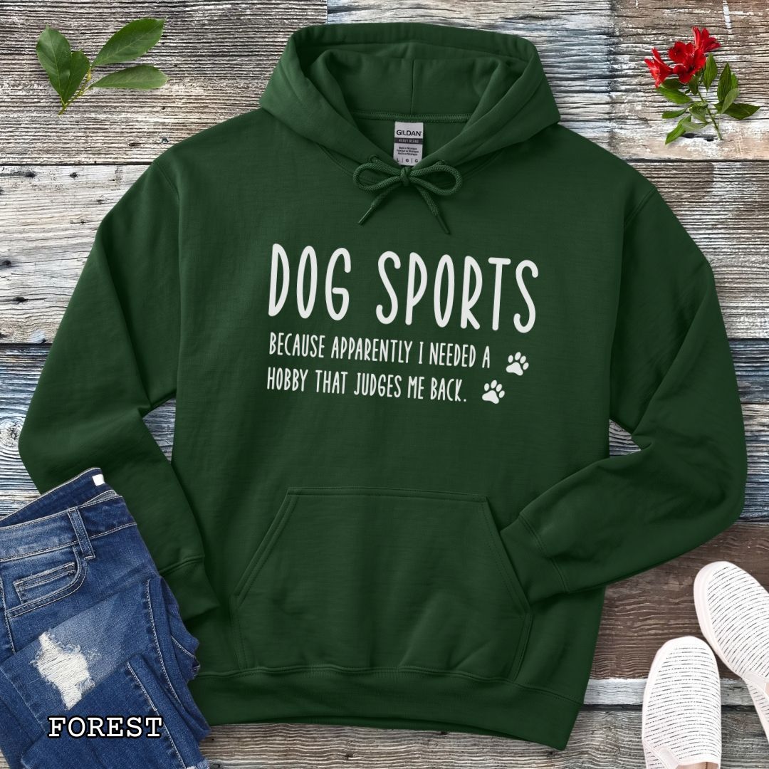 Dark green unisex hoodie for dog training enthusiasts with humorous text 'Dog Sports, Because Apparently I Needed a Hobby That Judges Me Back' accompanied by paw prints, shown on a wooden surface with jeans and flowers.