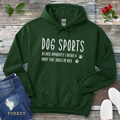 Dark green unisex hoodie for dog training enthusiasts with humorous text 'Dog Sports, Because Apparently I Needed a Hobby That Judges Me Back' accompanied by paw prints, shown on a wooden surface with jeans and flowers.