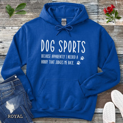 Royal blue unisex hoodie for dog training enthusiasts with humorous text 'Dog Sports, Because Apparently I Needed a Hobby That Judges Me Back' accompanied by paw prints, shown on a wooden surface with jeans and flowers.