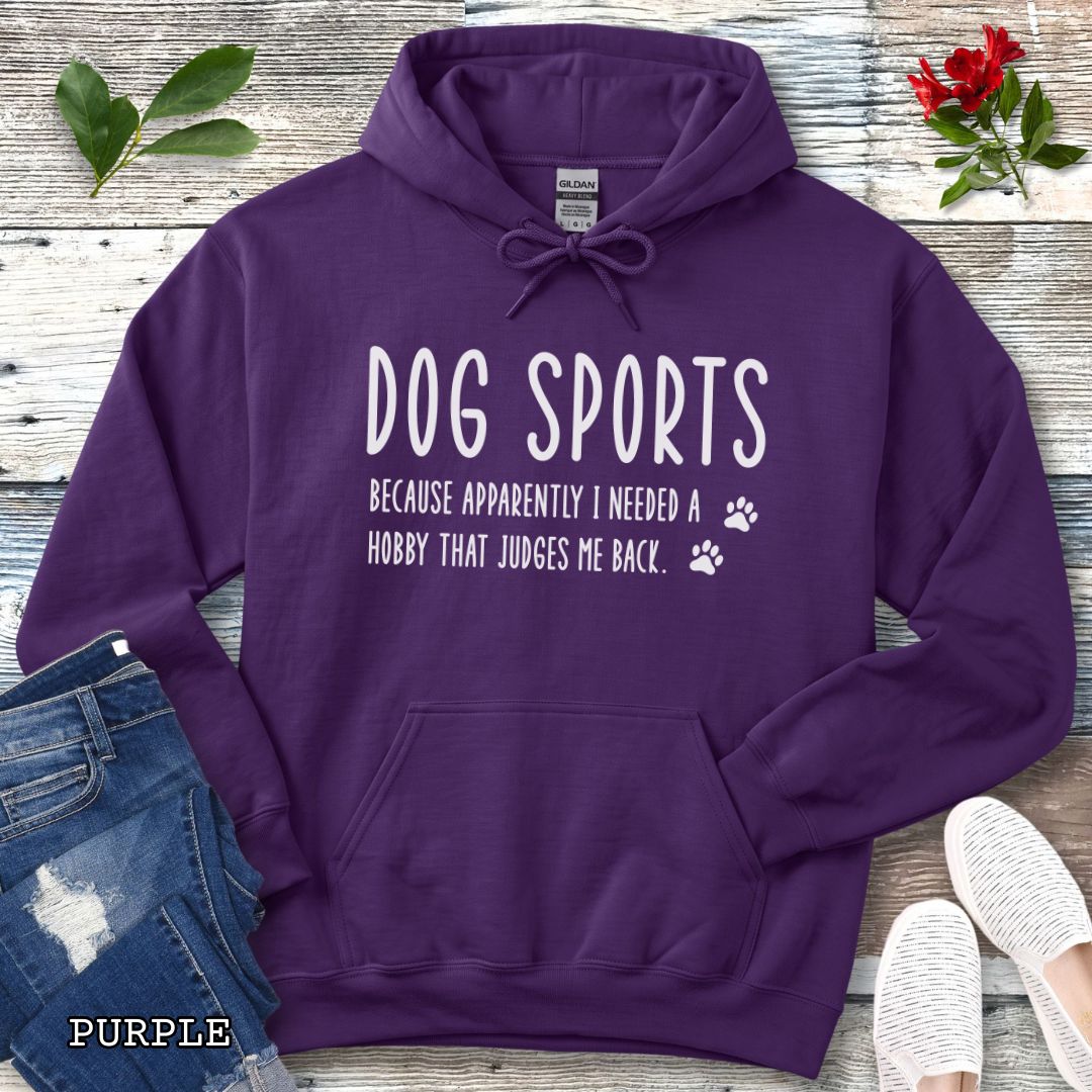 Purple unisex hoodie for dog training enthusiasts with humorous text 'Dog Sports, Because Apparently I Needed a Hobby That Judges Me Back' accompanied by paw prints, shown on a wooden surface with jeans and flowers.