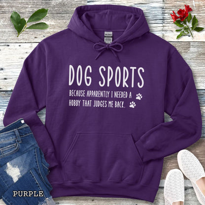 Purple unisex hoodie for dog training enthusiasts with humorous text 'Dog Sports, Because Apparently I Needed a Hobby That Judges Me Back' accompanied by paw prints, shown on a wooden surface with jeans and flowers.