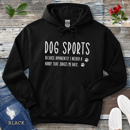 Black unisex hoodie for dog training enthusiasts with humorous text 'Dog Sports, Because Apparently I Needed a Hobby That Judges Me Back' accompanied by paw prints, shown on a wooden surface with jeans and flowers.