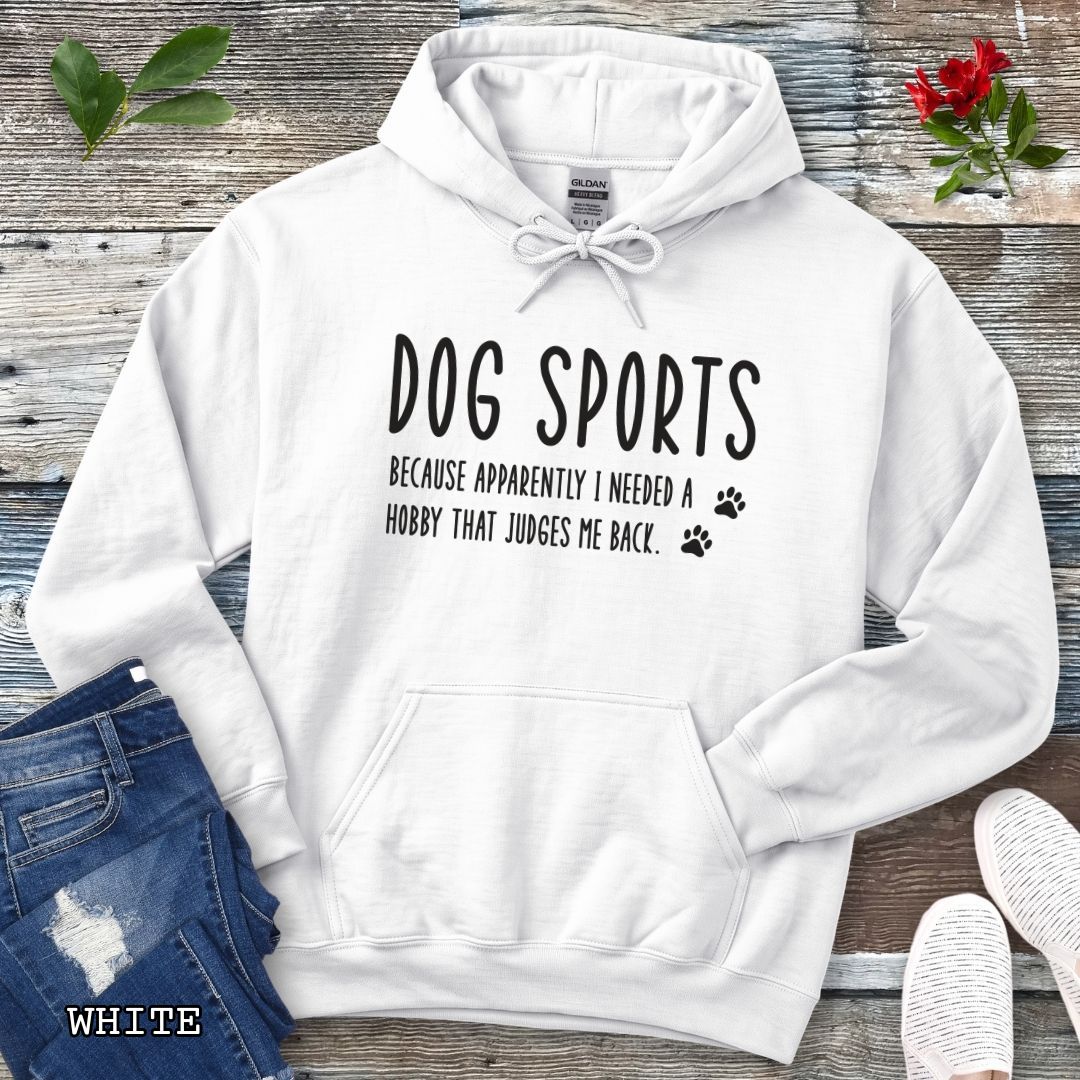 White unisex hoodie for dog training enthusiasts with humorous text 'Dog Sports, Because Apparently I Needed a Hobby That Judges Me Back' accompanied by paw prints, shown on a wooden surface with jeans and flowers.