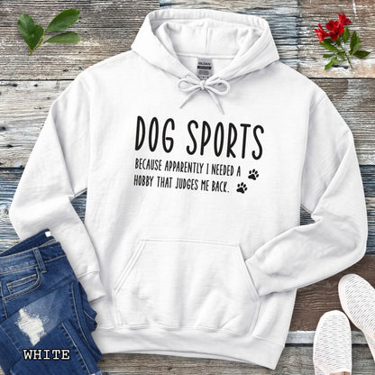 White unisex hoodie for dog training enthusiasts with humorous text 'Dog Sports, Because Apparently I Needed a Hobby That Judges Me Back' accompanied by paw prints, shown on a wooden surface with jeans and flowers.
