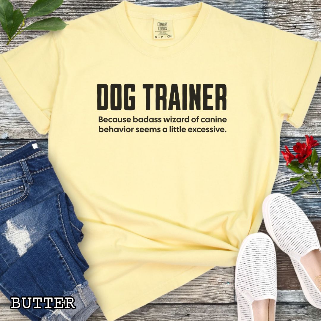 Yellow Comfort Colors t-shirt with humorous text 'DOG TRAINER, because badass wizard of canine behavior seems a little excessive', shown on a wooden surface with sunglasses and jeans.