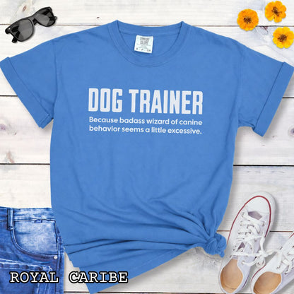 Royal blue Comfort Colors t-shirt with humorous text 'DOG TRAINER, because badass wizard of canine behavior seems a little excessive', shown on a wooden surface with sunglasses and jeans.