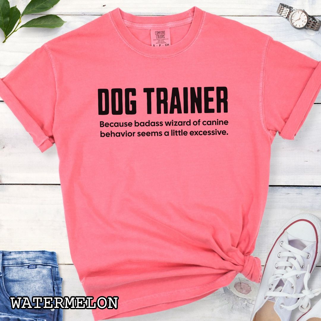 Coral pink Comfort Colors t-shirt with humorous text 'DOG TRAINER, because badass wizard of canine behavior seems a little excessive', shown on a wooden surface with sunglasses and jeans.