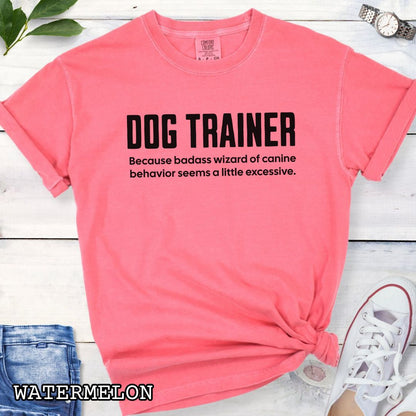Coral pink Comfort Colors t-shirt with humorous text 'DOG TRAINER, because badass wizard of canine behavior seems a little excessive', shown on a wooden surface with sunglasses and jeans.