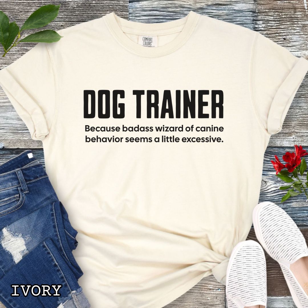 Beige Comfort Colors t-shirt with humorous text 'DOG TRAINER, because badass wizard of canine behavior seems a little excessive', shown on a wooden surface with sunglasses and jeans.