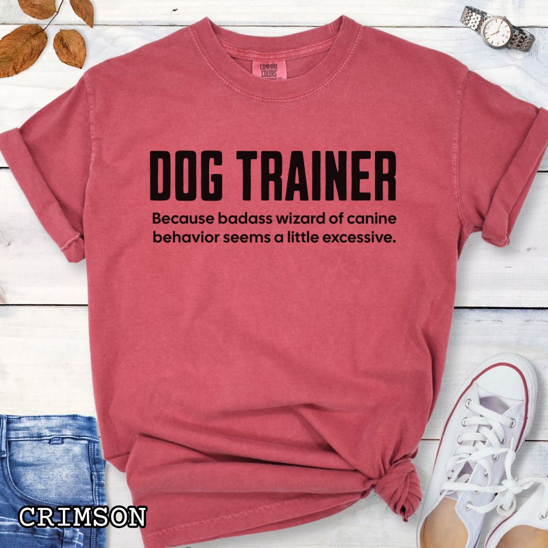 Red Comfort Colors t-shirt with humorous text 'DOG TRAINER, because badass wizard of canine behavior seems a little excessive', shown on a wooden surface with sunglasses and jeans.