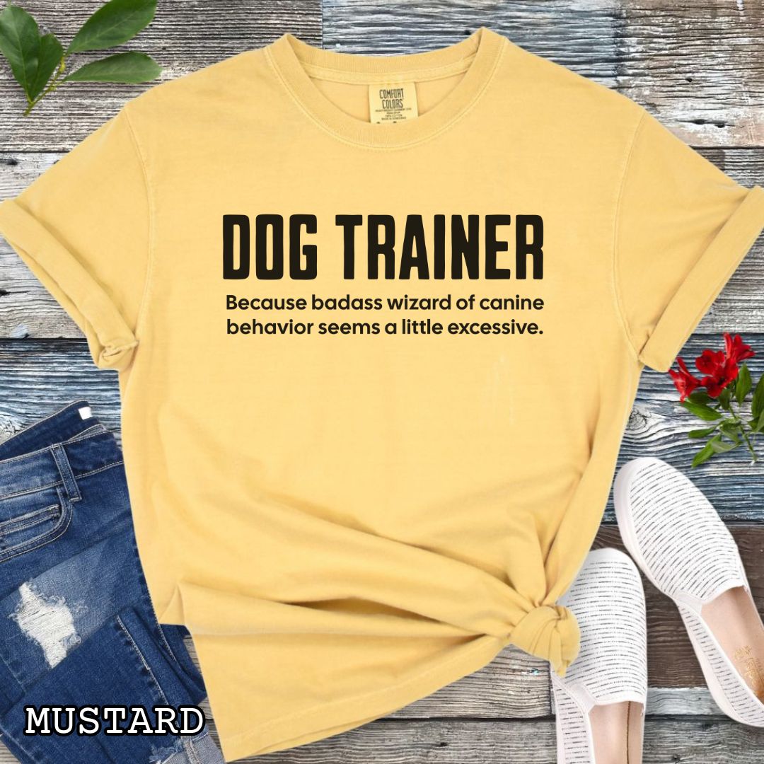 Mustard yellow Comfort Colors t-shirt with humorous text 'DOG TRAINER, because badass wizard of canine behavior seems a little excessive', shown on a wooden surface with sunglasses and jeans.
