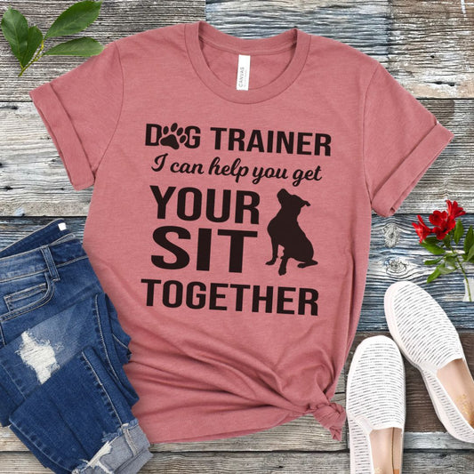 Mauve t-shirt with 'Dog Trainer, I can help you get your sit together' text and a sitting dog graphic, presented on a wooden surface with jeans and shoes.