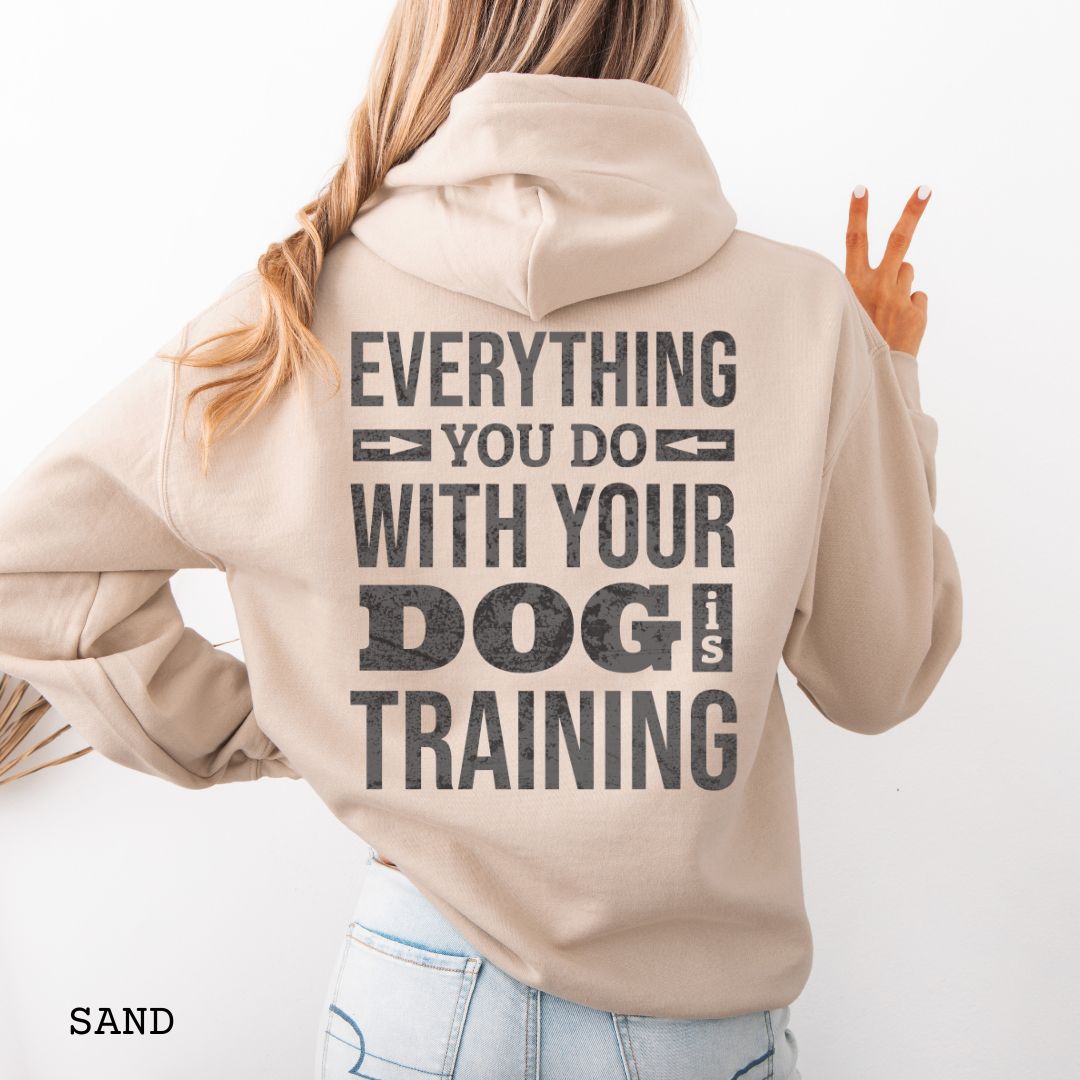 Person wearing a beige hoodie with the text 'EVERYTHING YOU DO WITH YOUR DOG IS TRAINING' text design printed on the back.