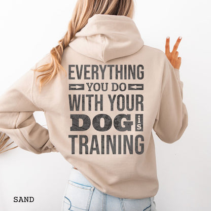 Person wearing a beige hoodie with the text 'EVERYTHING YOU DO WITH YOUR DOG IS TRAINING' text design printed on the back.