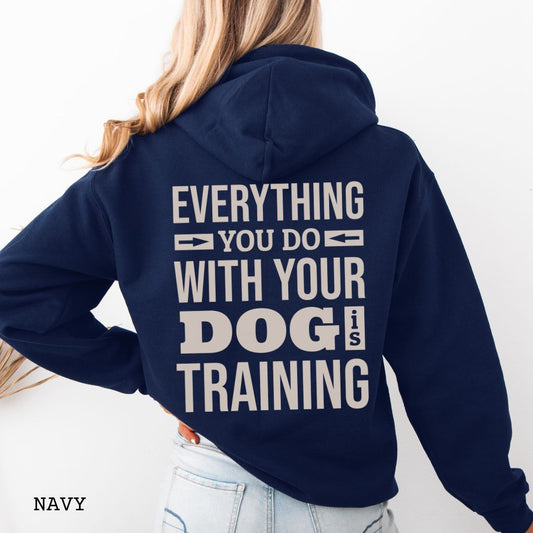 Person wearing a Navy blue hoodie with the text 'EVERYTHING YOU DO WITH YOUR DOG IS TRAINING' text design printed on the back.