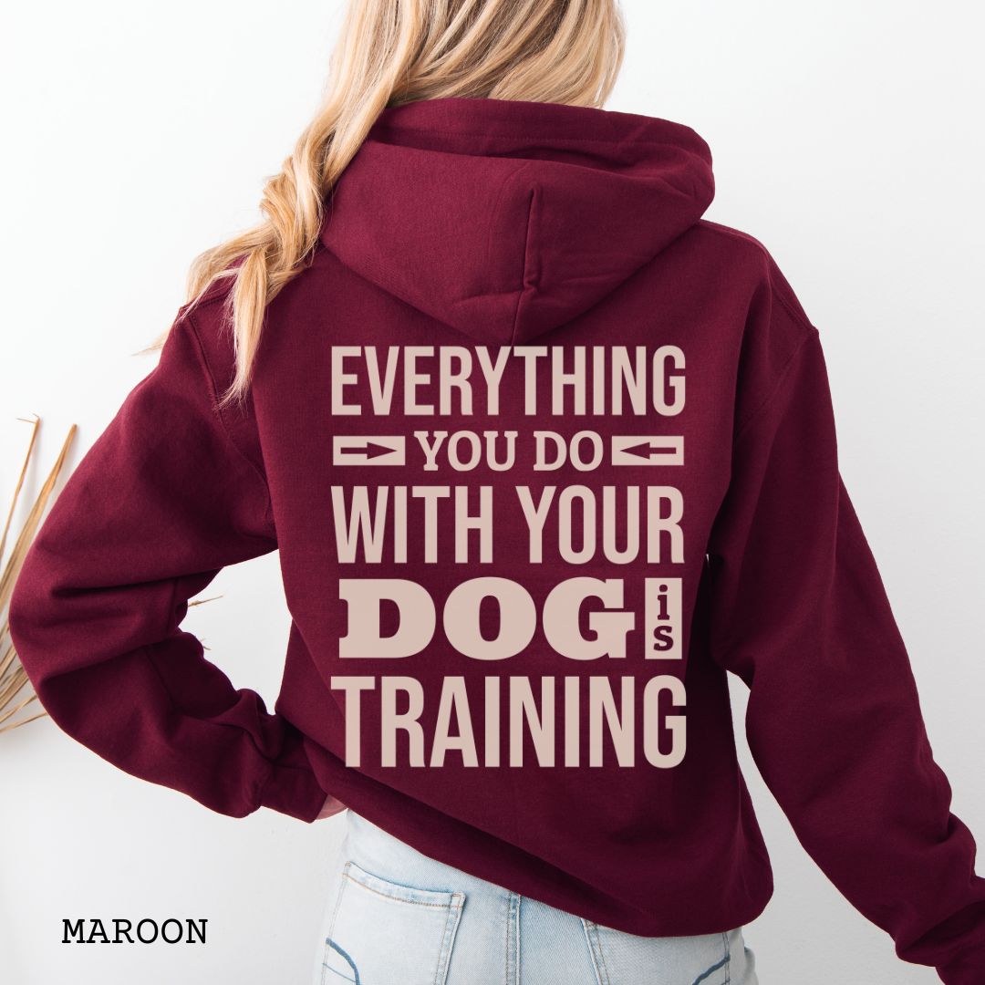 Person wearing a maroon hoodie with the text 'EVERYTHING YOU DO WITH YOUR DOG IS TRAINING' text design printed on the back.