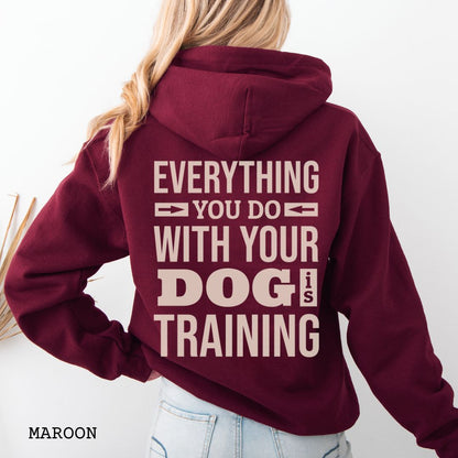 Person wearing a maroon hoodie with the text 'EVERYTHING YOU DO WITH YOUR DOG IS TRAINING' text design printed on the back.