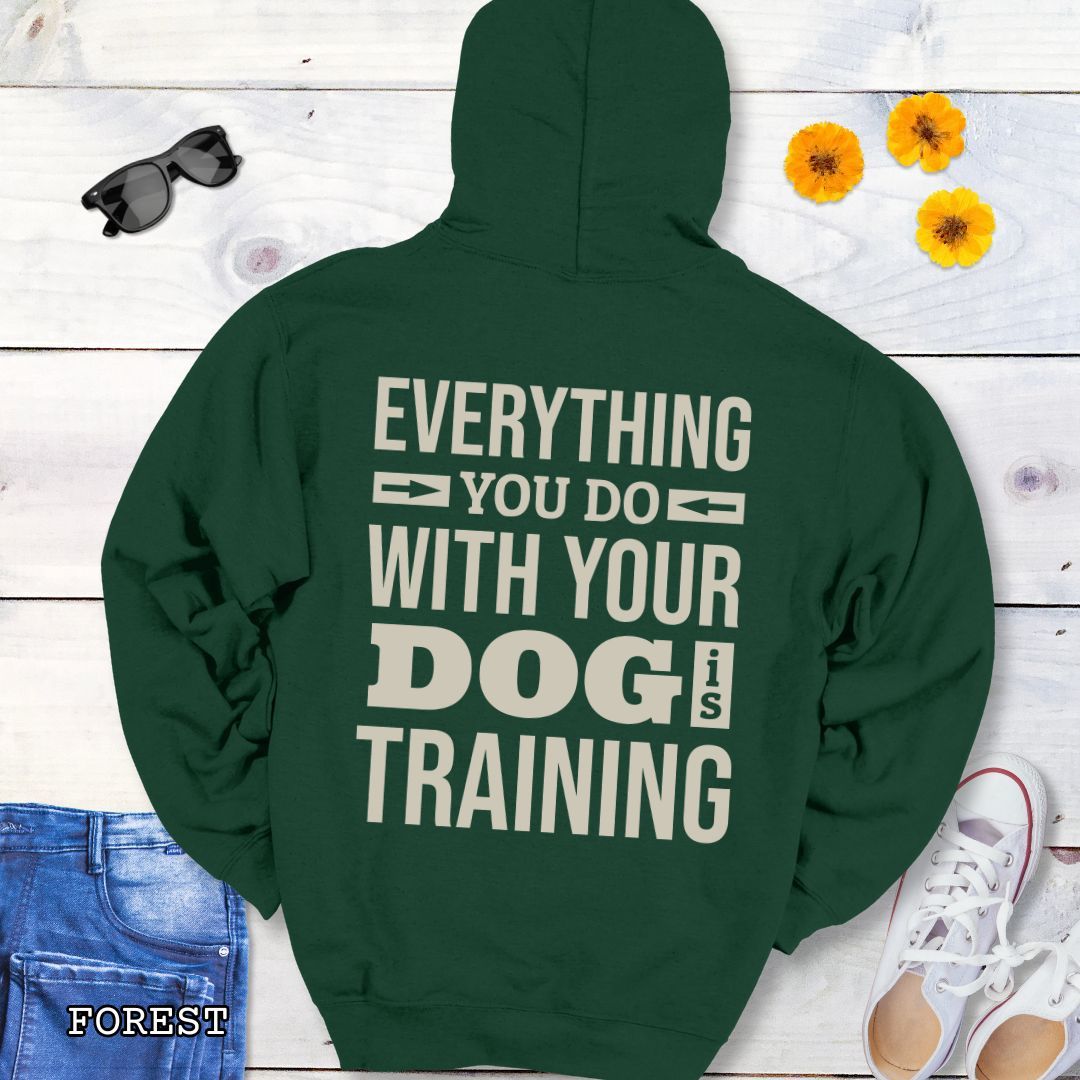 Dark green hoodie with the text 'EVERYTHING YOU DO WITH YOUR DOG IS TRAINING' text design printed on the back, displayed flat with a pair of blue jeans and white shoes.