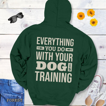 Dark green hoodie with the text 'EVERYTHING YOU DO WITH YOUR DOG IS TRAINING' text design printed on the back, displayed flat with a pair of blue jeans and white shoes.