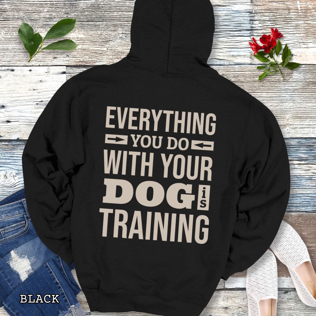 Black hoodie with the text 'EVERYTHING YOU DO WITH YOUR DOG IS TRAINING' text design printed on the back, displayed flat with a pair of blue jeans and white shoes.
