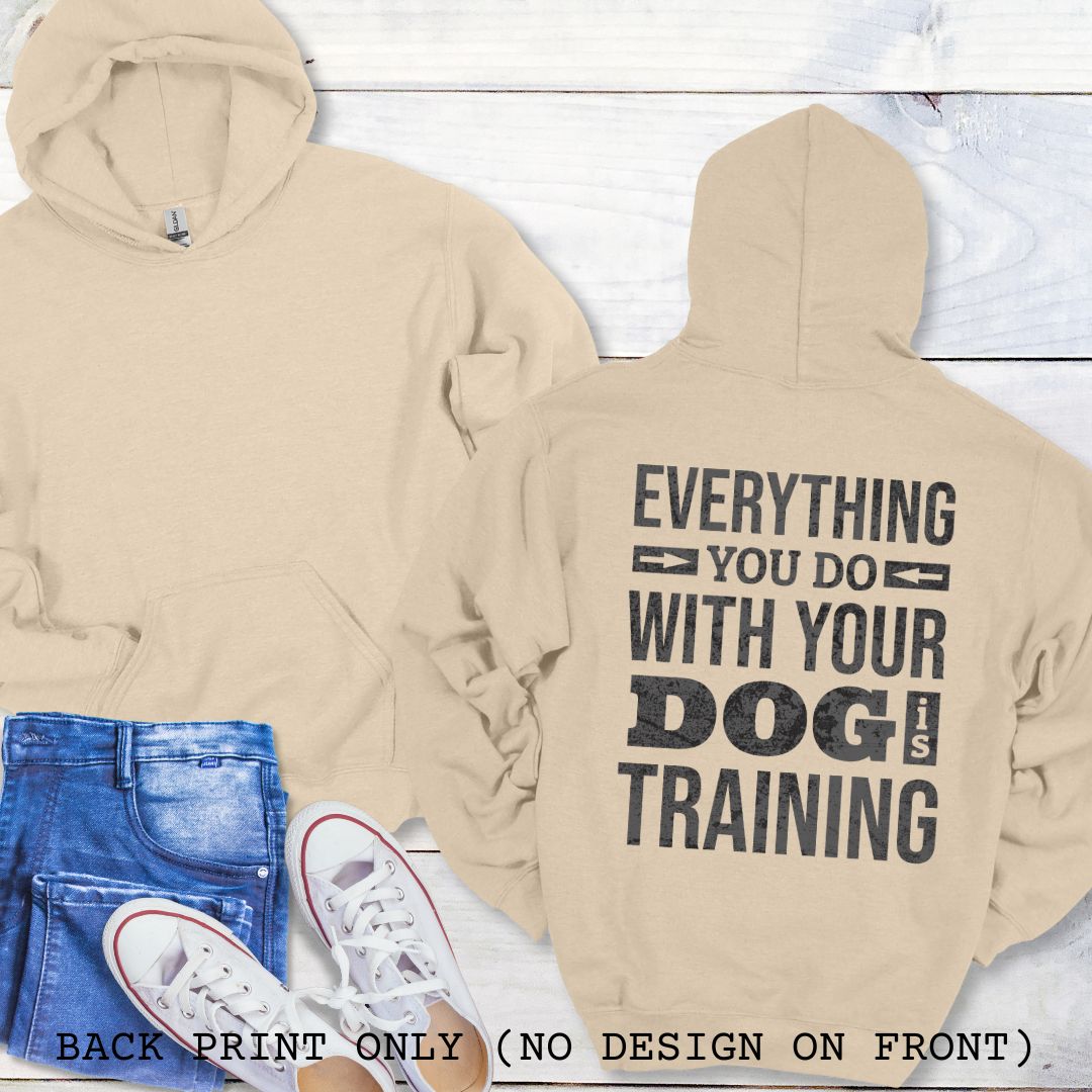 A sand colored hoodie with the text 'EVERYTHING YOU DO WITH YOUR DOG IS TRAINING' text design printed on the back, displayed flat with a pair of blue jeans and white shoes.