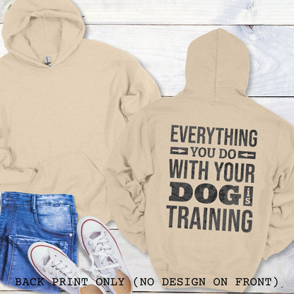 A sand colored hoodie with the text 'EVERYTHING YOU DO WITH YOUR DOG IS TRAINING' text design printed on the back, displayed flat with a pair of blue jeans and white shoes.