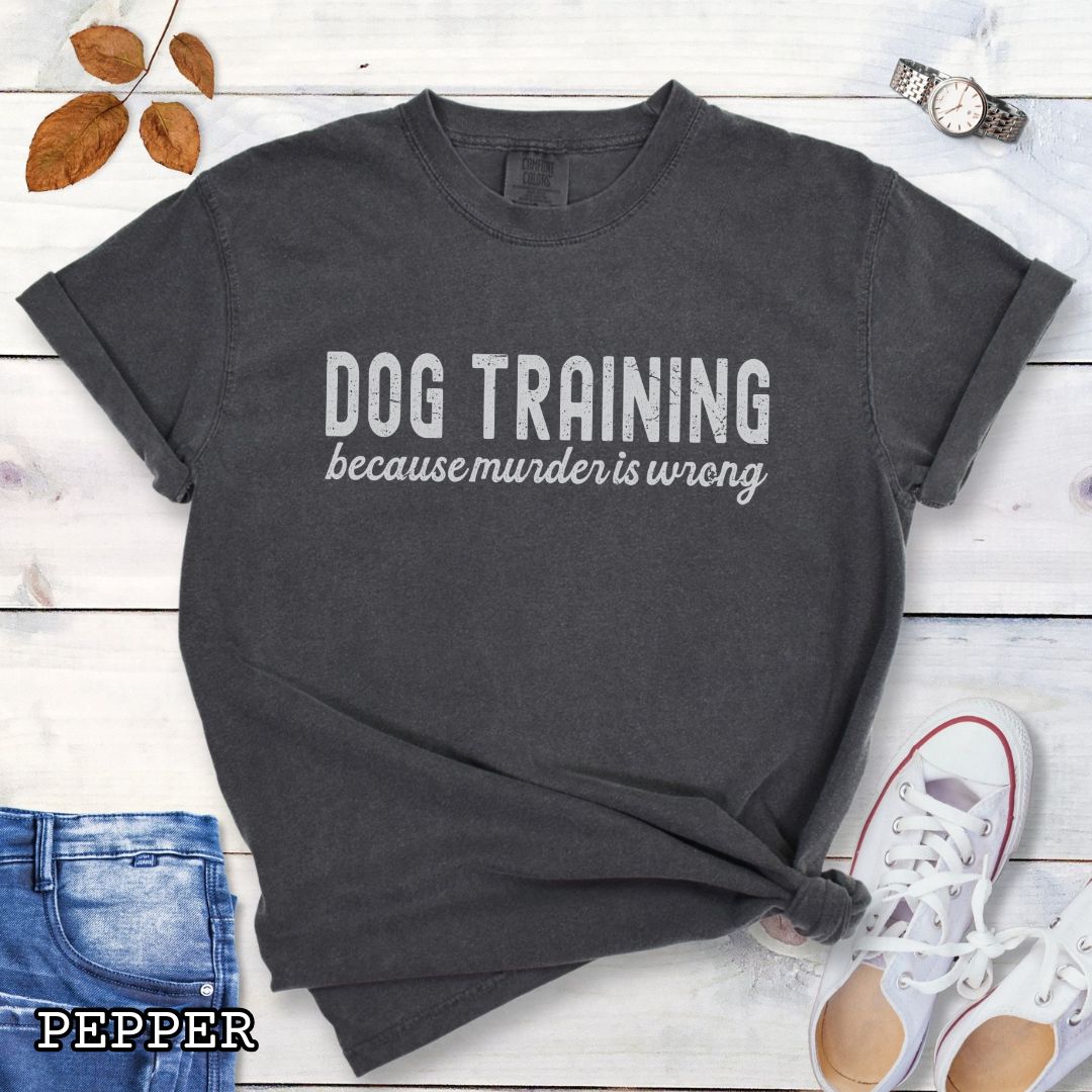 Grey (Pepper) Comfort Colors t-shirt with 'Dog Training because murder is wrong' text on a wooden surface with sunglasses, jeans, and sneakers.