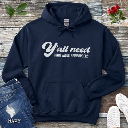 A navy blue pullover hoodie for reward-based dog trainers with humorous text 'Y'all need HIGH VALUE REINFORCERS' across the chest in retro lettering, styled in a flat-lay arrangement with blue jeans, white shoes, and small green and red floral accents on a rustic wood background.