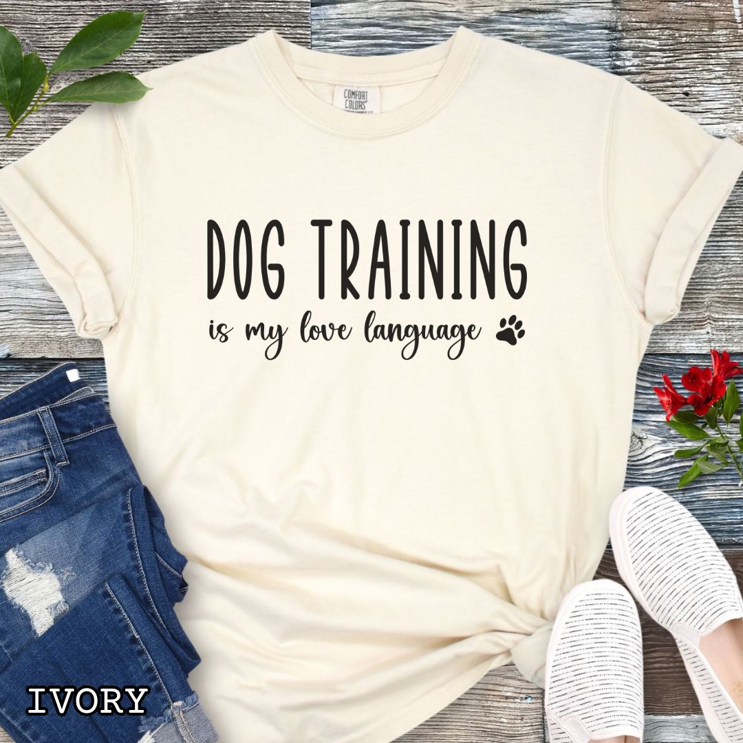 Beige (Ivory) Comfort Colors t-shirt with 'Dog training is my love language' text and a small paw print, presented on a wooden surface with jeans and shoes.