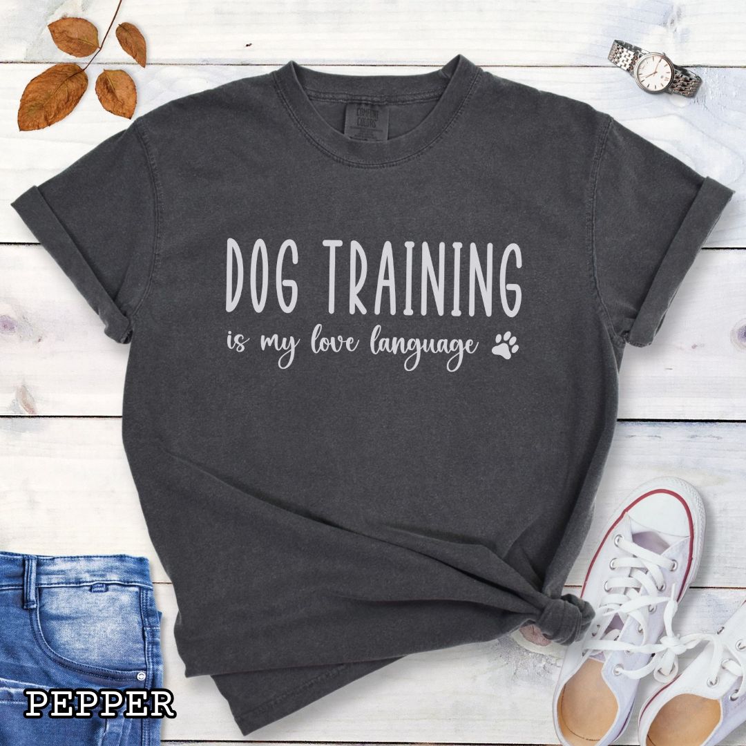Dark grey (Pepper) Comfort Colors t-shirt with 'Dog training is my love language' text and a small paw print, presented on a wooden surface with jeans and shoes.