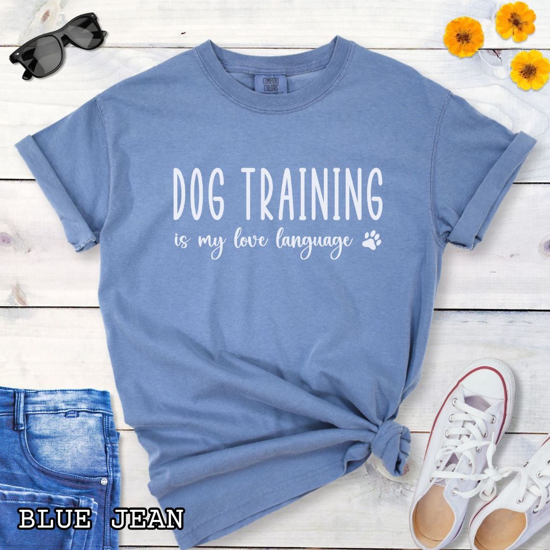 Blue Jean color Comfort Colors t-shirt with 'Dog training is my love language' text and a small paw print, presented on a wooden surface with jeans and shoes.