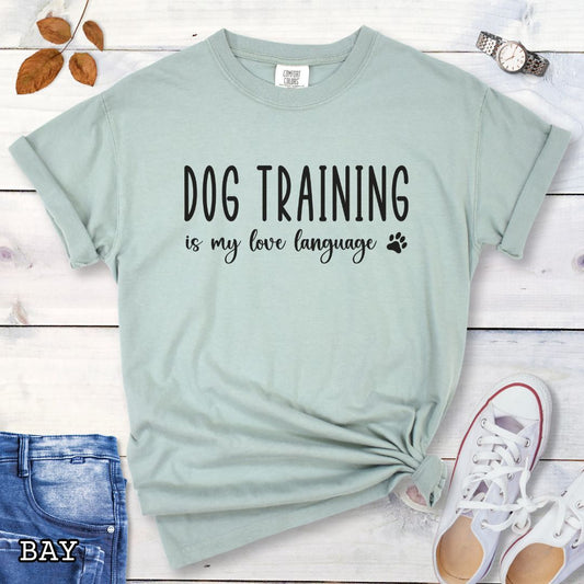 Light green (Bay) Comfort Colors t-shirt with 'Dog training is my love language' text and a small paw print, presented on a wooden surface with jeans and shoes.