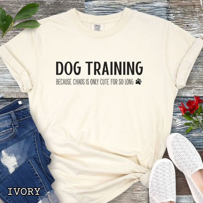 Beige (Ivory) Comfort Colors t-shirt with 'Dog Training, Because Chaos is Only Cute For So Long' text and a small paw print, on a wooden surface with jeans and sneakers.