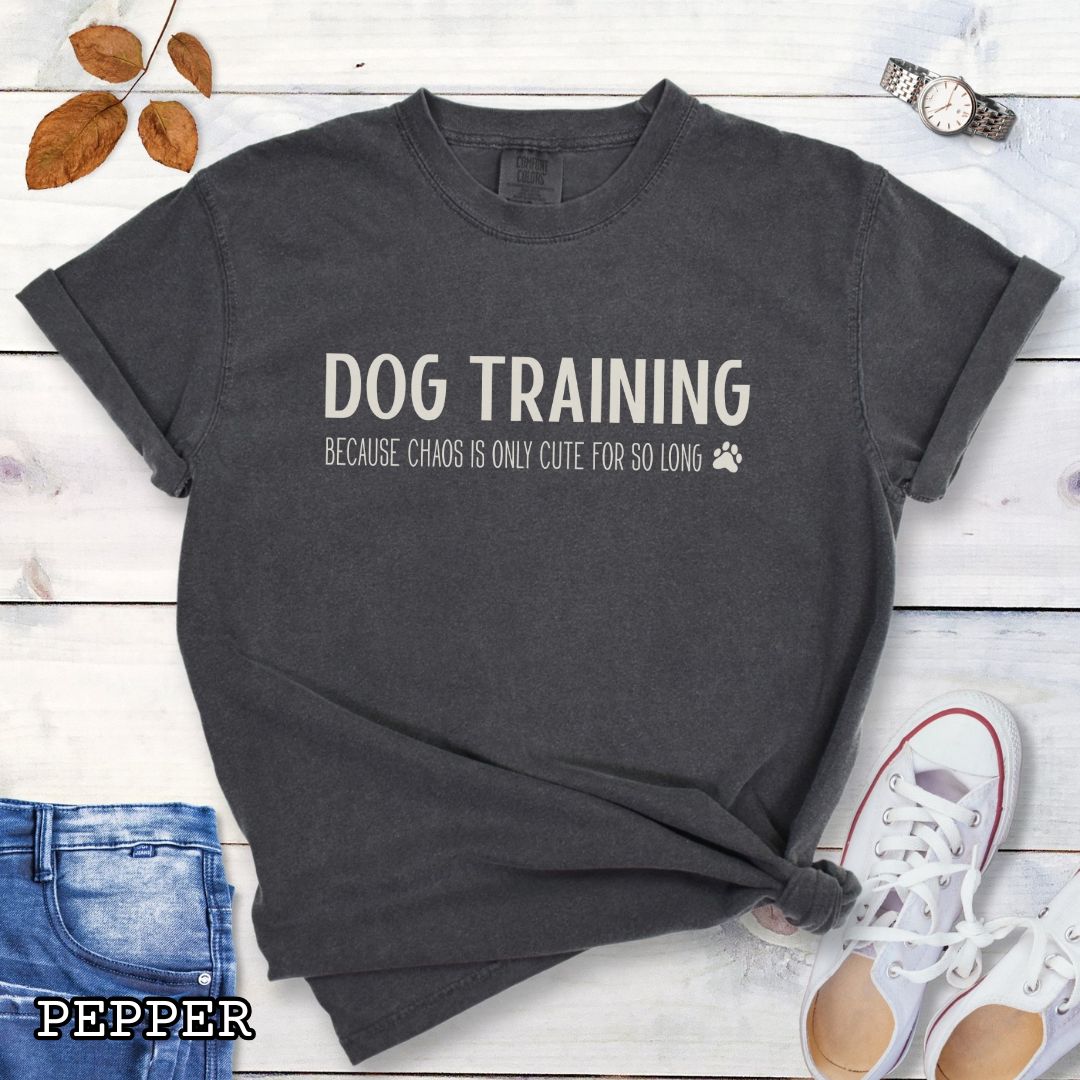 Dark grey (Pepper) Comfort Colors t-shirt with 'Dog Training, Because Chaos is Only Cute For So Long' text and a small paw print, on a wooden surface with jeans and sneakers.