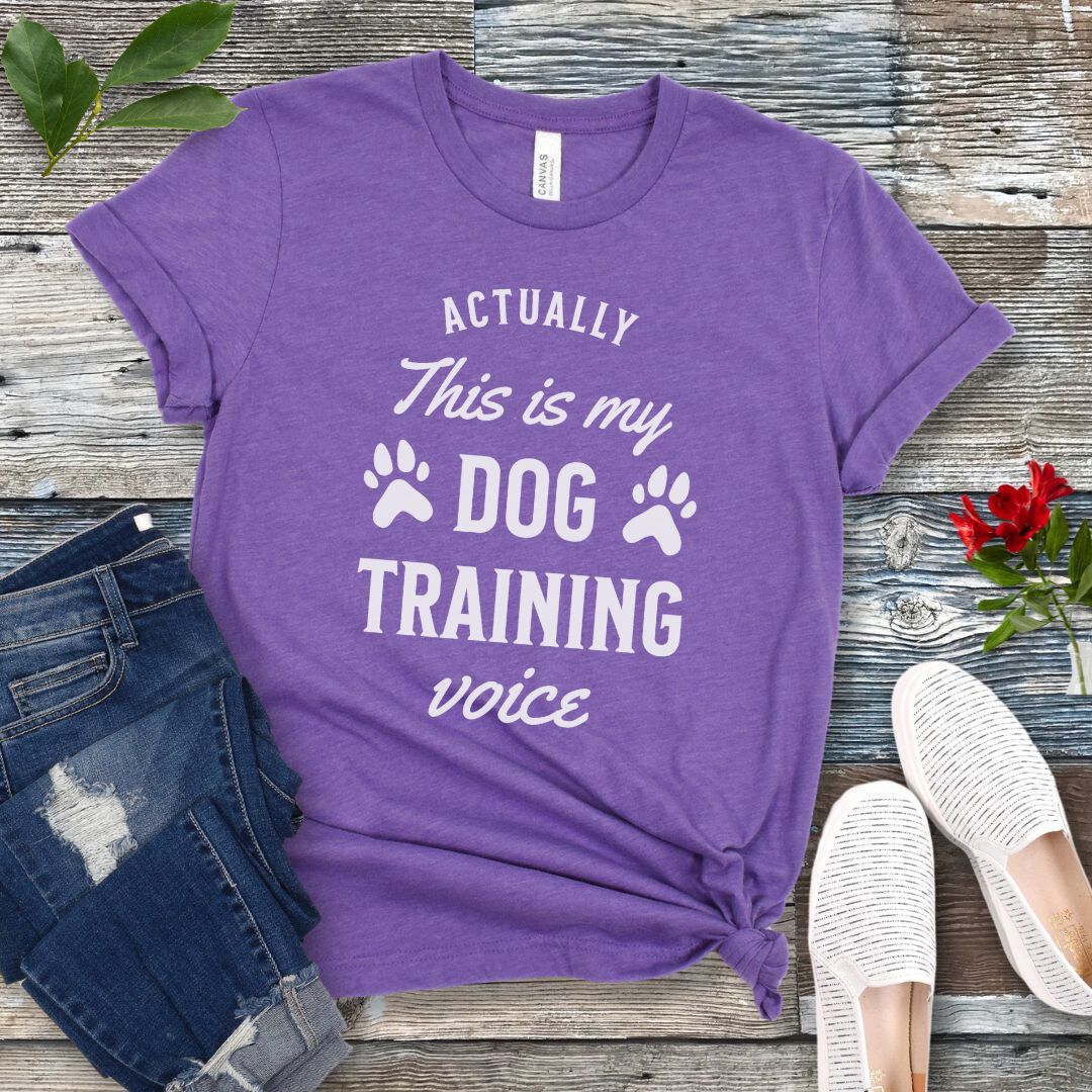 Purple t-shirt with 'Actually this is my dog training voice' text on a wooden surface with jeans and shoes.