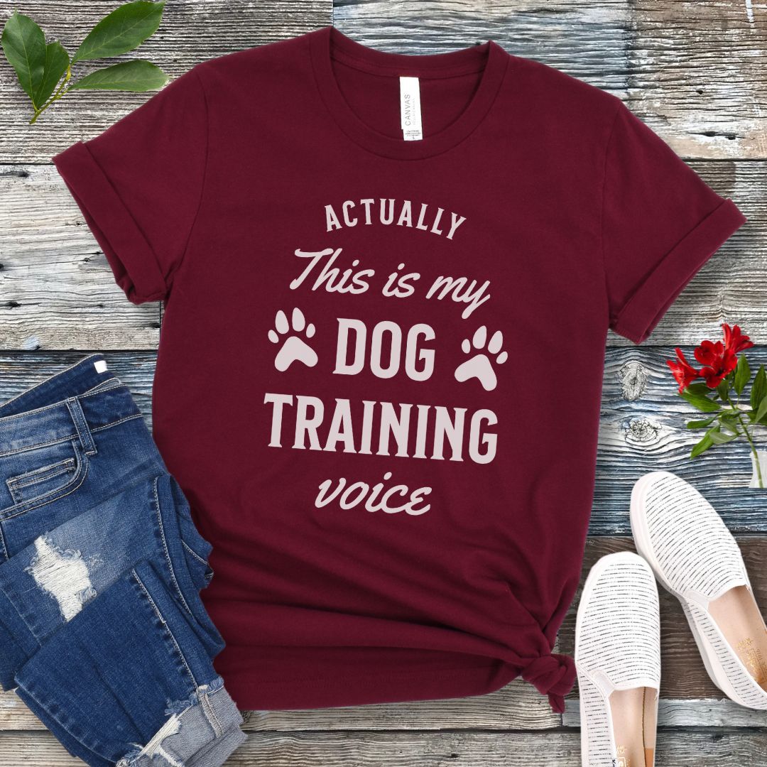 Maroon t-shirt with 'Actually this is my dog training voice' text on a wooden surface with jeans and shoes.
