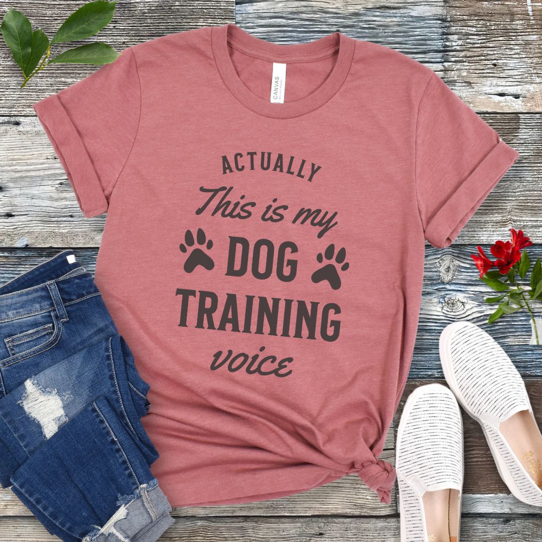 Mauve t-shirt with 'Actually this is my dog training voice' text on a wooden surface with jeans and shoes.
