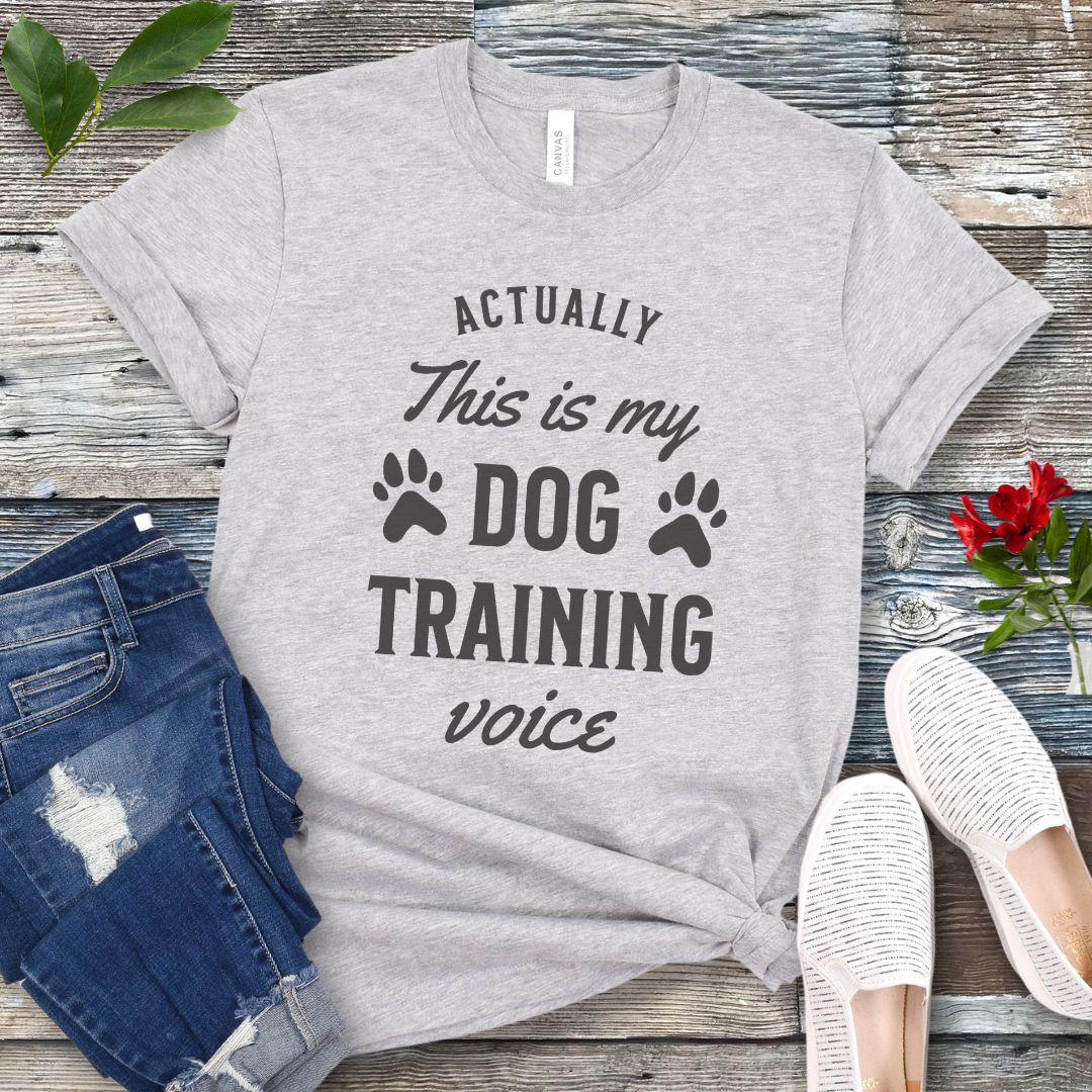 Light gray t-shirt with 'Actually this is my dog training voice' text on a wooden surface with jeans and shoes.