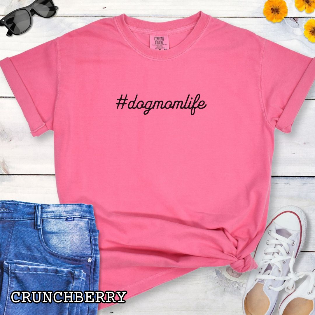 Pink Comfort Colors t-shirt for dog lovers with '#dogmomlife' text, shown on a wooden surface with shoes and jeans. 